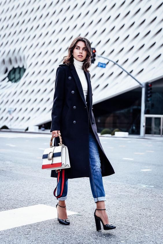 Contemporary winter outfit featuring a tailored black wool blend coat, white turtleneck, distressed jeans, and chic black heels in an urban setting.
