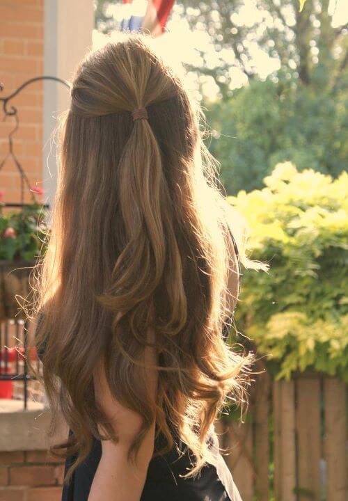 Long, flowing hair styled in a half-up bun, with soft waves framing the face. Natural lighting highlights the shiny texture against lush greenery.