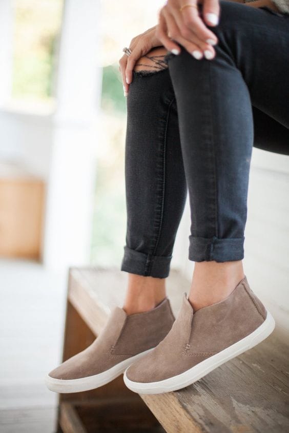 Light taupe suede sneaker shoes with a slip-on design and white rubber soles, paired with rolled-up dark denim jeans in a bright interior.