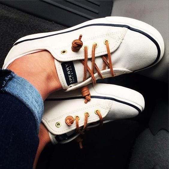 White low-top sneakers with navy blue piping, brown leather laces, paired with rolled-up jeans, embodying a casual, laid-back style.