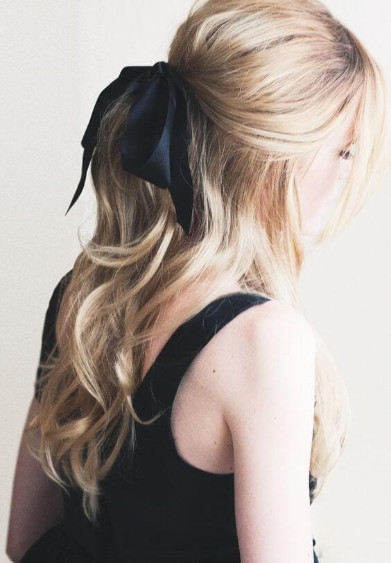 Long blonde hair styled in a half-up, half-down bun with soft waves and a black silk ribbon bow, perfect for formal occasions.