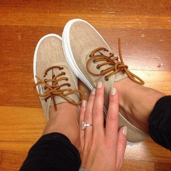 Beige canvas sneakers with brown laces and white rubber soles, styled on a wooden floor with a hand showcasing manicured nails.