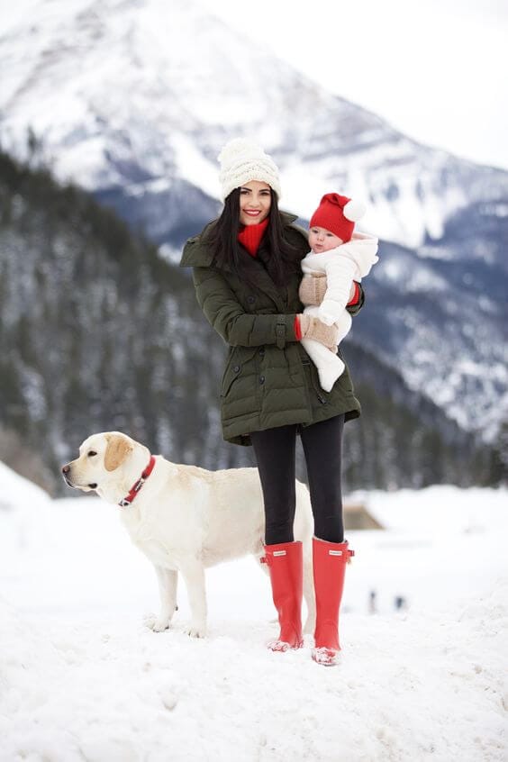 A woman in a dark green parka with red accents cradles a baby in a cream outfit against a snowy backdrop, wearing black leggings and red boots.