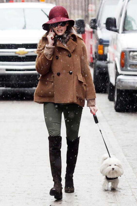 A woman walks on a snow-covered street in a chic brown double-breasted coat, olive leggings, and thigh-high black boots, holding a leash for a small white dog.