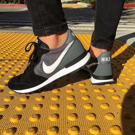 Black suede and mesh sneakers with a white logo, sturdy gray heel cap, styled on a yellow crosswalk mat, perfect for casual wear.