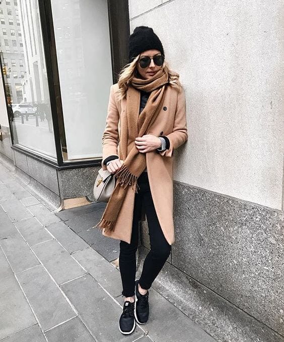 Chic winter outfit featuring a mid-thigh beige coat over a black top, skinny black pants, a neutral scarf, and stylish sneakers, perfect for cold weather.