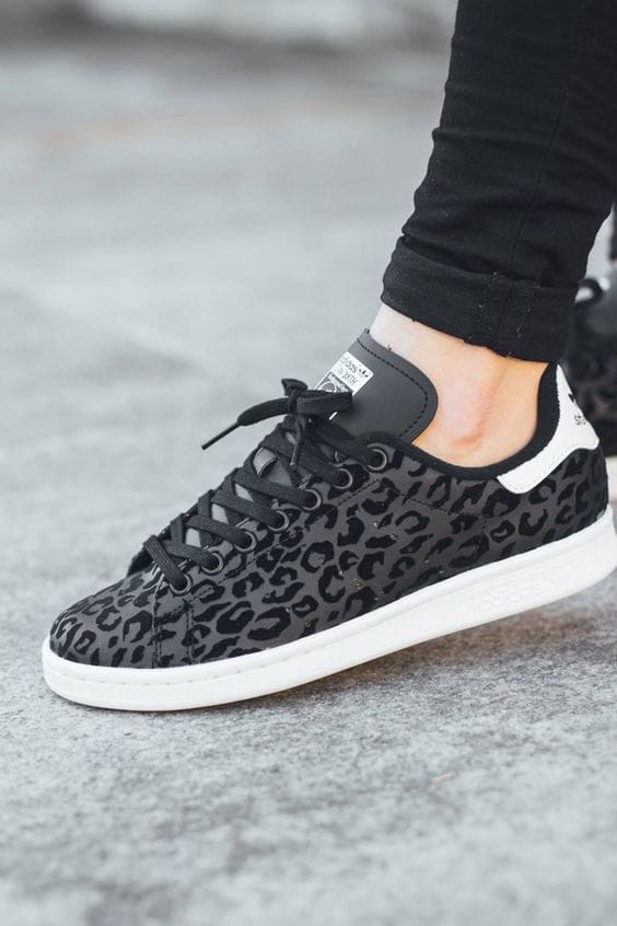 Sneakers with a black leopard print, lightweight for casual wear, featuring a white heel accent and flat rubber sole, styled with slim black trousers.