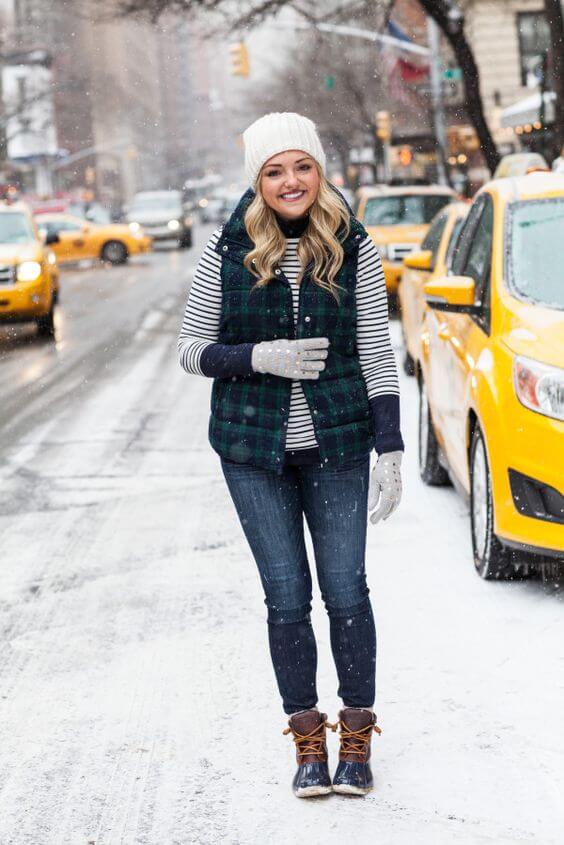 A young woman in a plaid puffer vest and striped shirt stands on a snowy NYC street, showcasing chic winter outfit ideas.