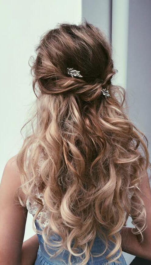 Hairstyle with cascading waves and soft curls, featuring a half-up bun secured by decorative hairpins in warm blonde tones.
