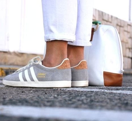 Sneakers with a grey suede upper and white leather stripes, paired with white cropped trousers, set against a stone background.