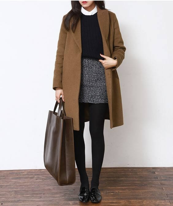 Camel coat layered over a navy sweater, paired with a black and white textured skirt and black tights. A brown leather tote complements the look.