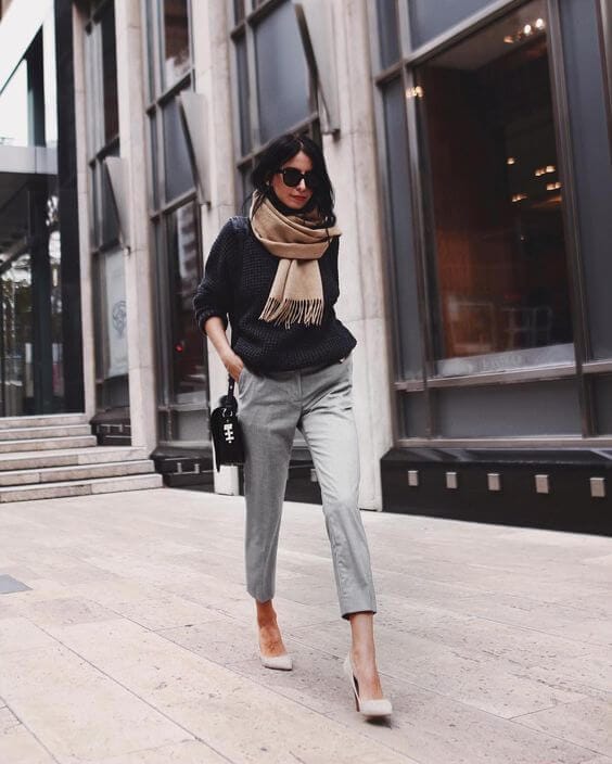 Model in a dark knit sweater and light grey tapered trousers, accessorized with a beige scarf and nude high heels, exuding winter office style.