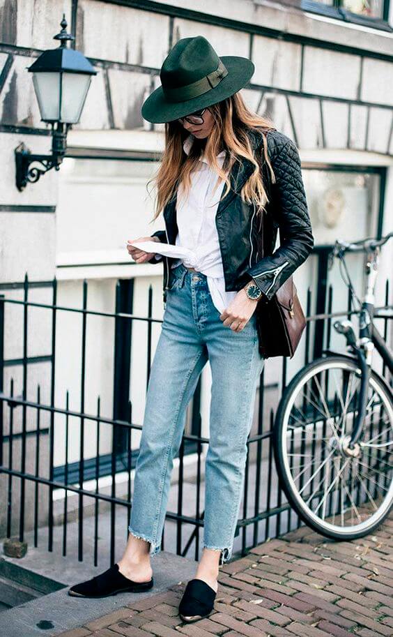 Stylish wide-brimmed green hat on flowing hair, paired with a black leather jacket, white shirt, and light blue jeans, exuding chic urban fashion.