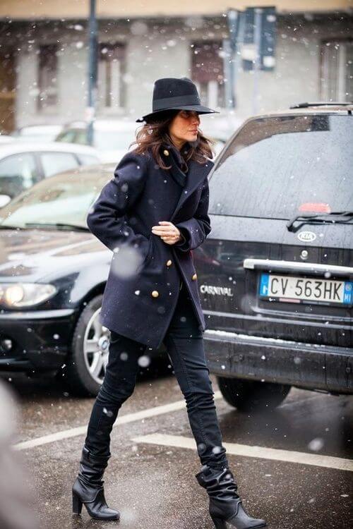 A woman in a snowy urban scene wears a long navy coat, fitted black pants, high-heeled boots, and a wide-brimmed black hat.