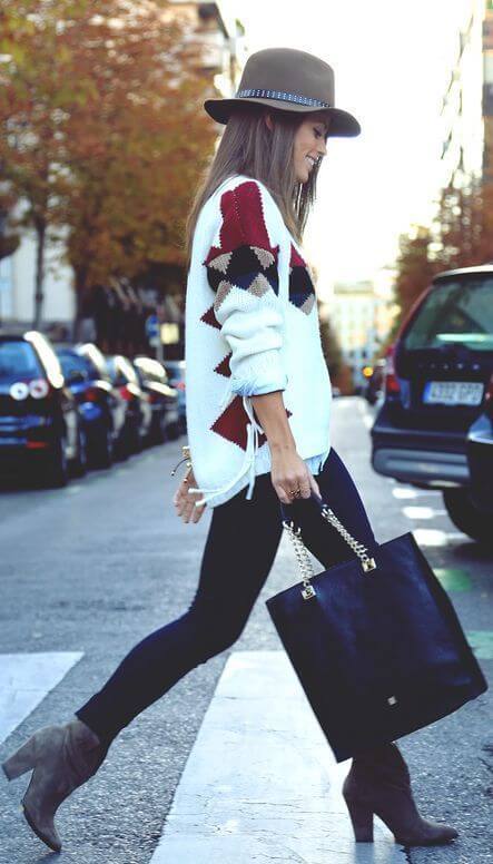 A woman in a white sweater and brown wide-brimmed hat stands on an urban street with autumn trees, showcasing winter fashion.