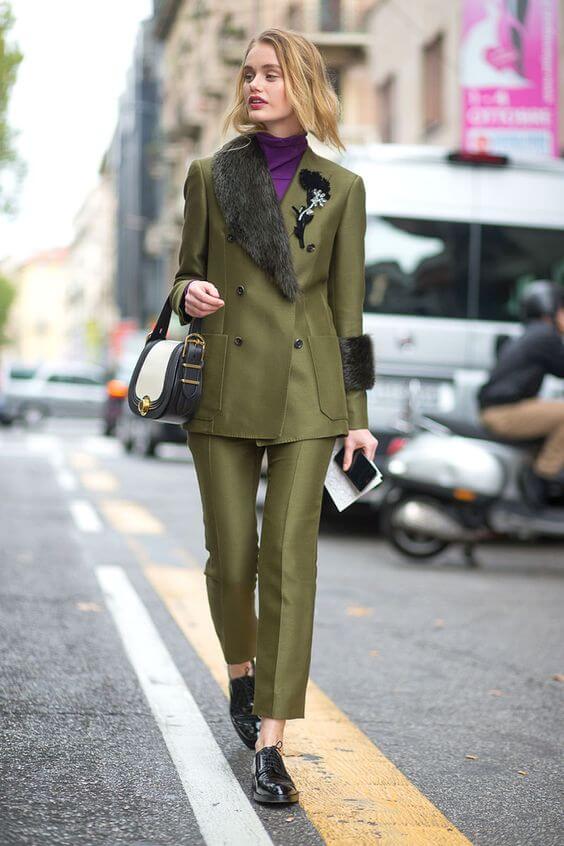 Tailored olive green suit with a double-breasted jacket, fur collar, floral brooch, slim-fit trousers, and black patent lace-up shoes.