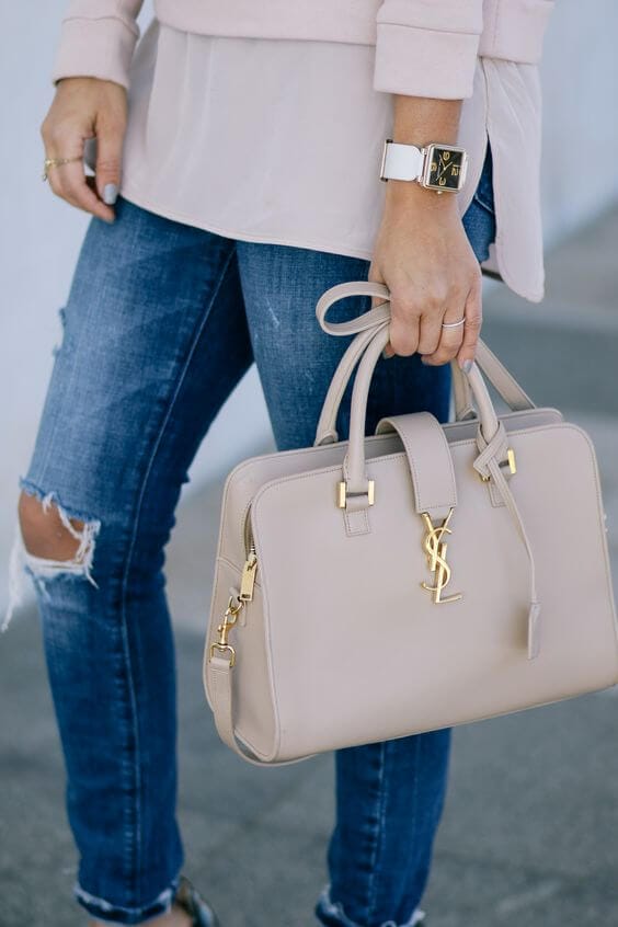 Structured beige leather handbag with gold-tone hardware and logo plaque, paired with a light pink top and distressed denim.