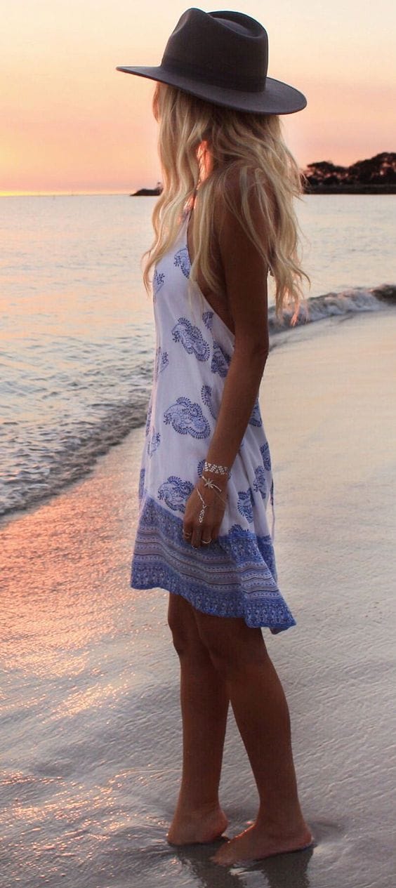 Wide-brimmed gray straw hat complements a flowing blue and white paisley dress, perfect for beach outings at sunset.