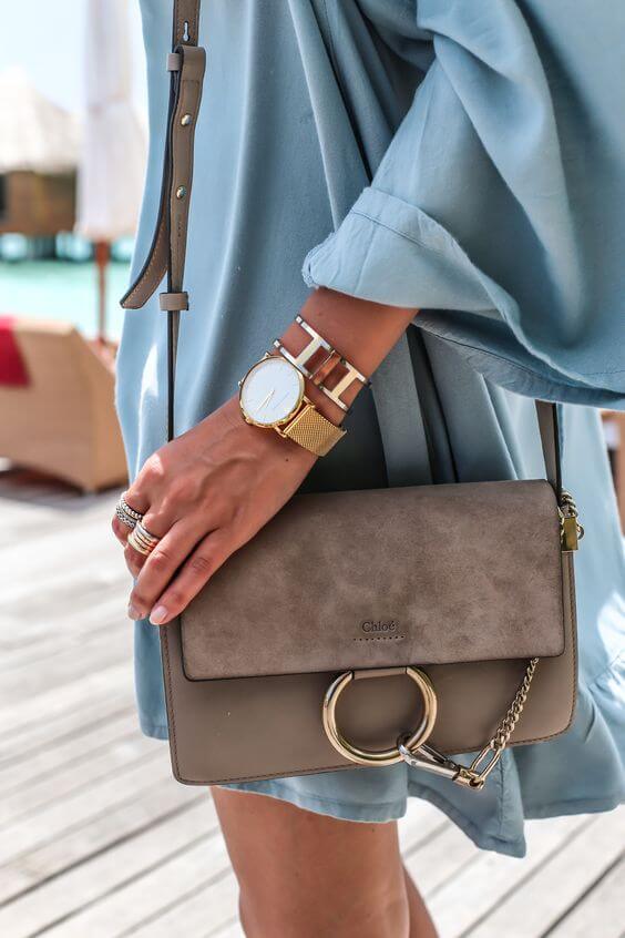 Chloé handbag in taupe suede with structured design, circular handle, and detachable chain strap. Paired with a light blue garment and gold watch.