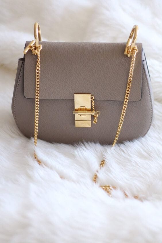 Taupe leather handbag with a structured silhouette, gold lock closure, and detachable chain strap, set on a soft white fur background.