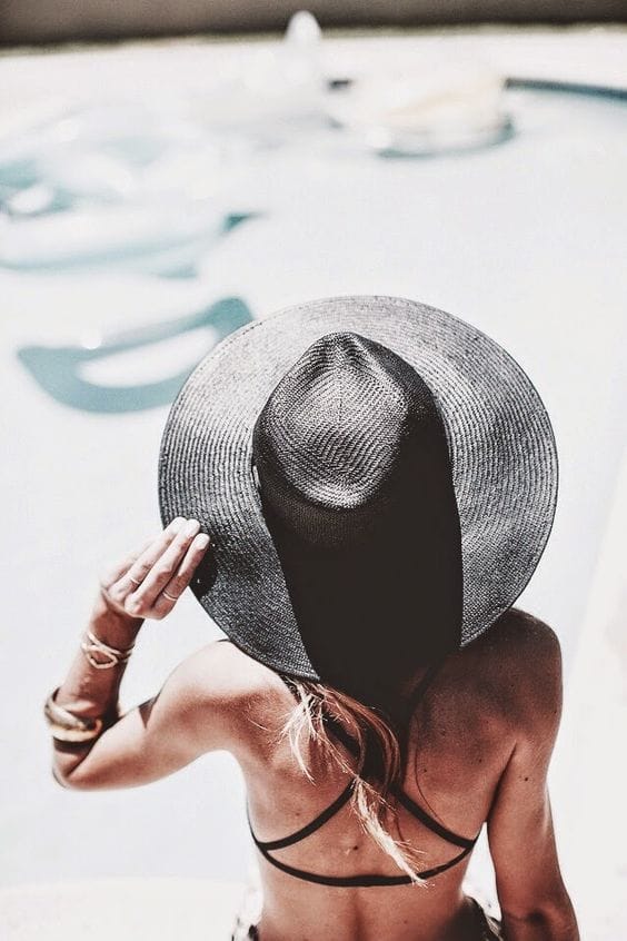 Wide-brimmed woven straw beach hat in classic black and natural colors, worn poolside by a person with long, light brown hair.