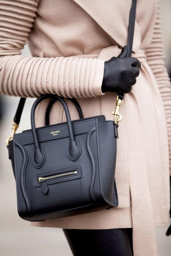 Structured black leather handbag with zip compartment, double handles, and detachable strap, worn with a ribbed beige coat in an urban setting.