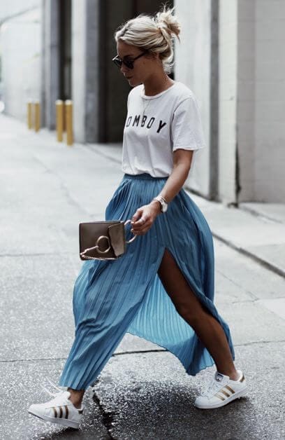 Casual outfit featuring a white t-shirt with bold print, flowing blue maxi skirt with high side slit, and white sporty sneakers. Urban setting.