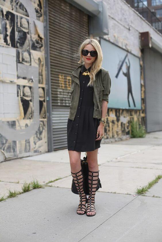 Layered outfit featuring a black dress and olive green utility jacket, paired with knee-high black gladiator sandals and chunky jewelry.