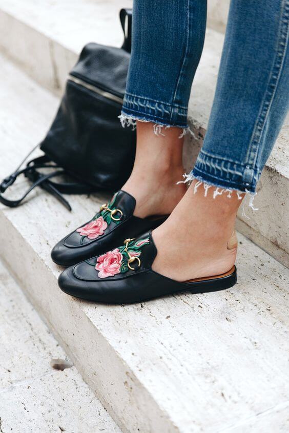 Sleek black leather mules with floral embroidery paired with distressed blue jeans, sitting on a stone step, embodying summer fashion.
