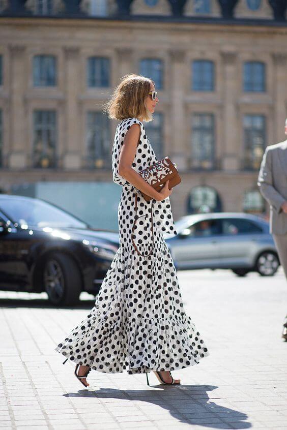 Polka dot dress in white with large black dots; sleeveless, flared silhouette, ruffled hem. Paired with brown handbag and black heels, urban backdrop.