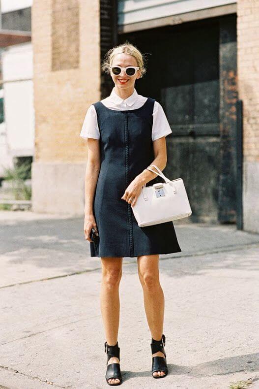 Sleeveless black dress with a fitted silhouette, white collared shirt underneath, paired with a small white handbag and black heeled sandals.