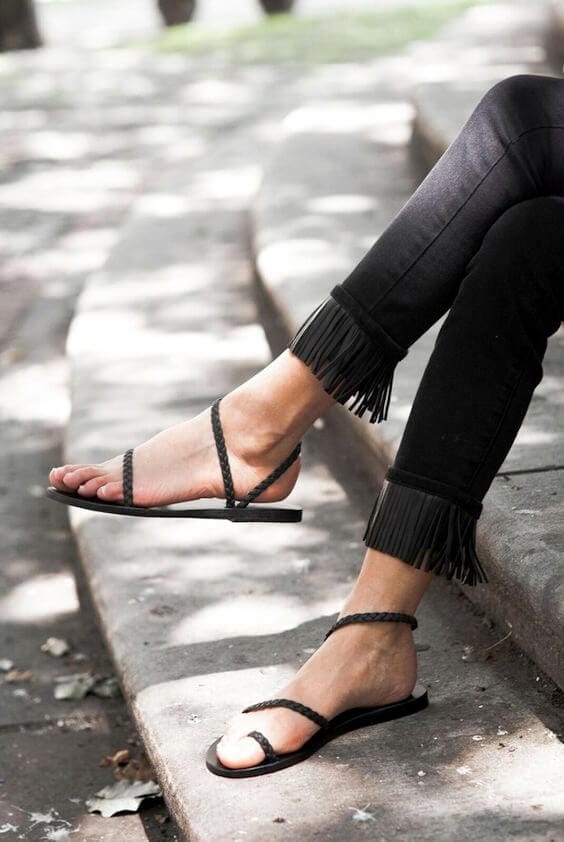 Black flat sandals with thin straps paired with fringe-detailed black skinny jeans, set outdoors in dappled sunlight, ideal for summer outfits.