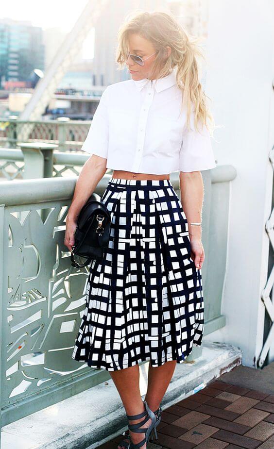 Cropped white button-up shirt paired with a high-waisted black and white grid-patterned skirt, strappy gray heels, and a black handbag.