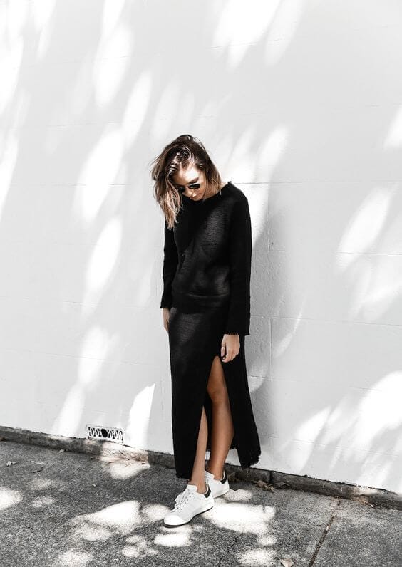 A woman in a black, long-sleeved knitted top and high-slit black skirt, paired with white sneakers, exudes urban fashion elegance.