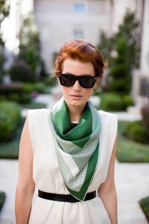 Oversized silk scarf with a geometric green and white pattern elegantly draped around the neck, paired with a sleeveless white dress, set against lush greenery.
