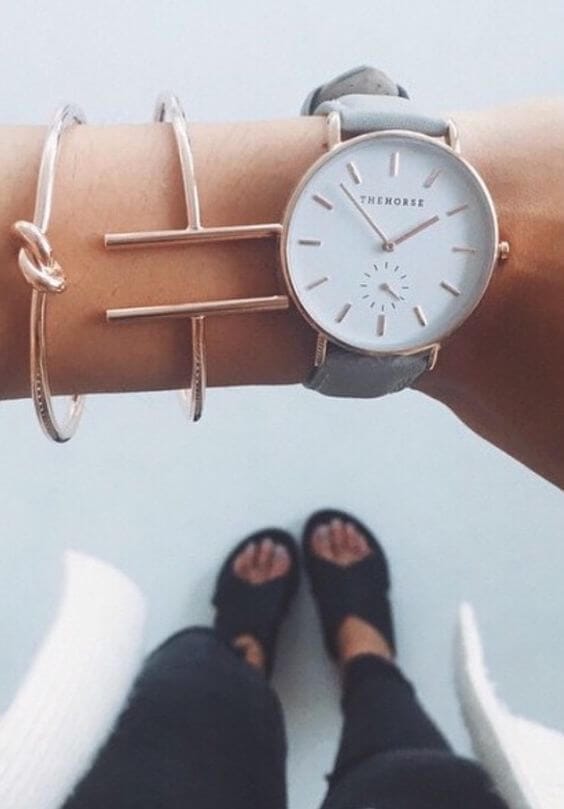 Minimalist fashion accessories featuring a silver-faced watch, grey leather strap, and two metallic bracelets, emphasizing stylish wrist adornments.