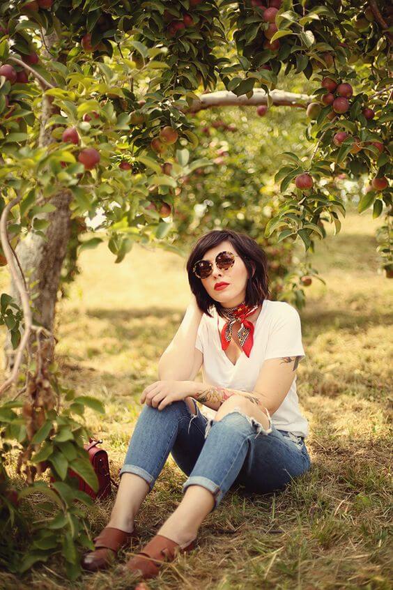 Medium-length, textured bob with soft waves, dark brown hair, vintage sunglasses, and a red scarf, seated under an apple tree.