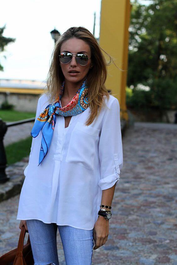 Vibrant silk scarf in blue and orange tied around the neck, paired with a white button-up shirt, light blue jeans, and oversized sunglasses.