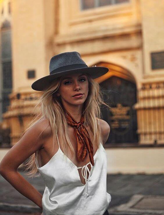Loose waves cascade past the shoulders, complemented by a silky white top and earthy toned scarf. A wide-brimmed gray hat enhances the chic summer look.
