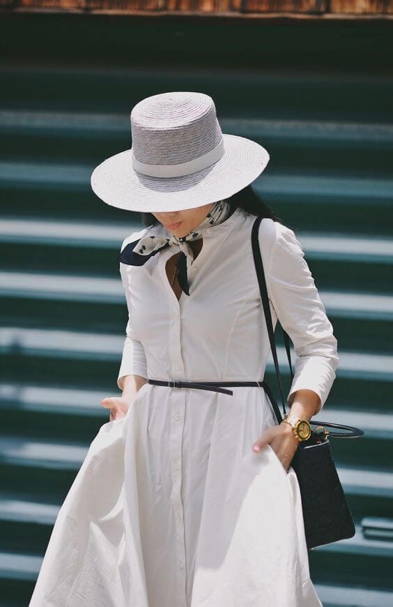 White knee-length shirt dress paired with a chic light scarf, gray straw hat, and small black handbag for summer elegance.