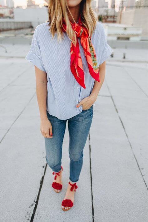 A light blue vertically striped blouse, fitted denim jeans, and a red silk scarf create a chic summer outfit, complemented by red heeled sandals.