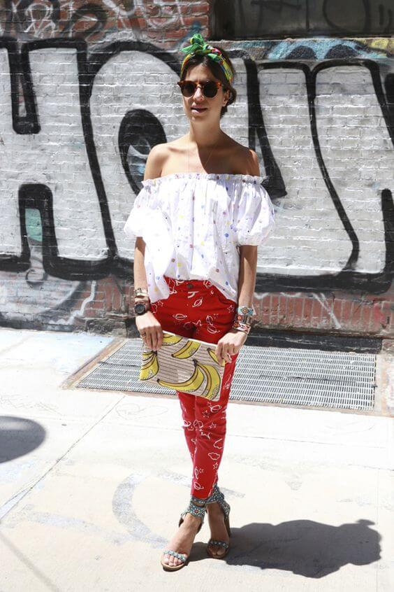 Off-the-shoulder white blouse with puffed sleeves, high-waisted red patterned pants, layered bracelets, and a colorful headscarf.