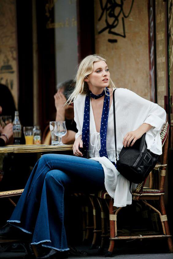 A model sits at a cafe in a flowy white top and high-waisted flared jeans, styled with a navy polka dot scarf, exuding chic summer fashion.