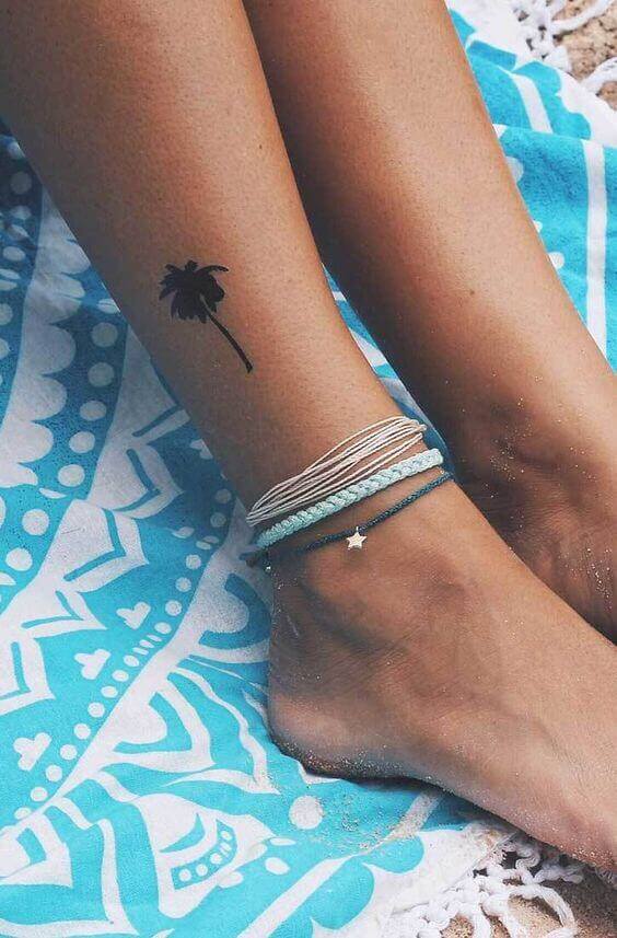 Tropical palm tree tattoo on inner ankle, surrounded by layered boho ankle bracelets, set against a vibrant blue patterned towel.