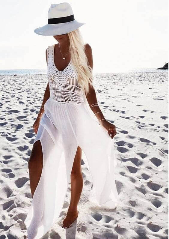 Flowing white maxi dress with crochet top and side slits, paired with a wide-brimmed hat, perfect for summer beach outings.