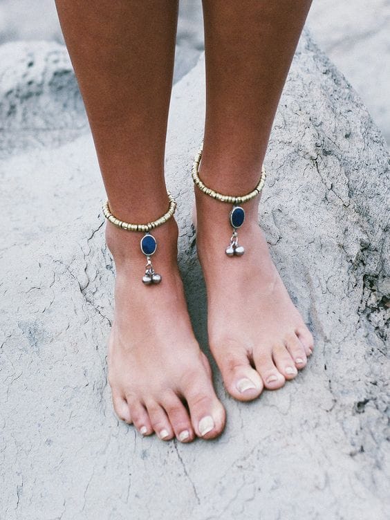 Anklet with golden beads and a blue gem, designed for boho style. Accentuates bare feet on rocky surface, perfect for beach settings.