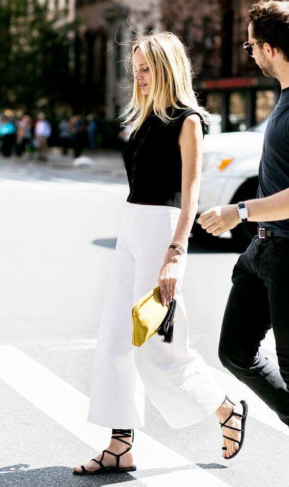 Confident woman in a fitted black sleeveless top and wide-legged white trousers, accessorized with a yellow clutch, walking on a city street.