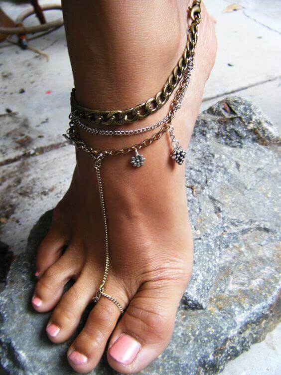 Foot jewelry features layered gold and silver-toned chains around the ankle, adorned with charms, complementing light pink nail polish.