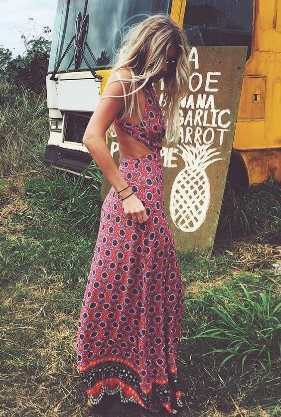 A woman in a vibrant, flowing dress with a halter neckline poses in tall grass near a rustic bus, evoking a relaxed beach vibe.