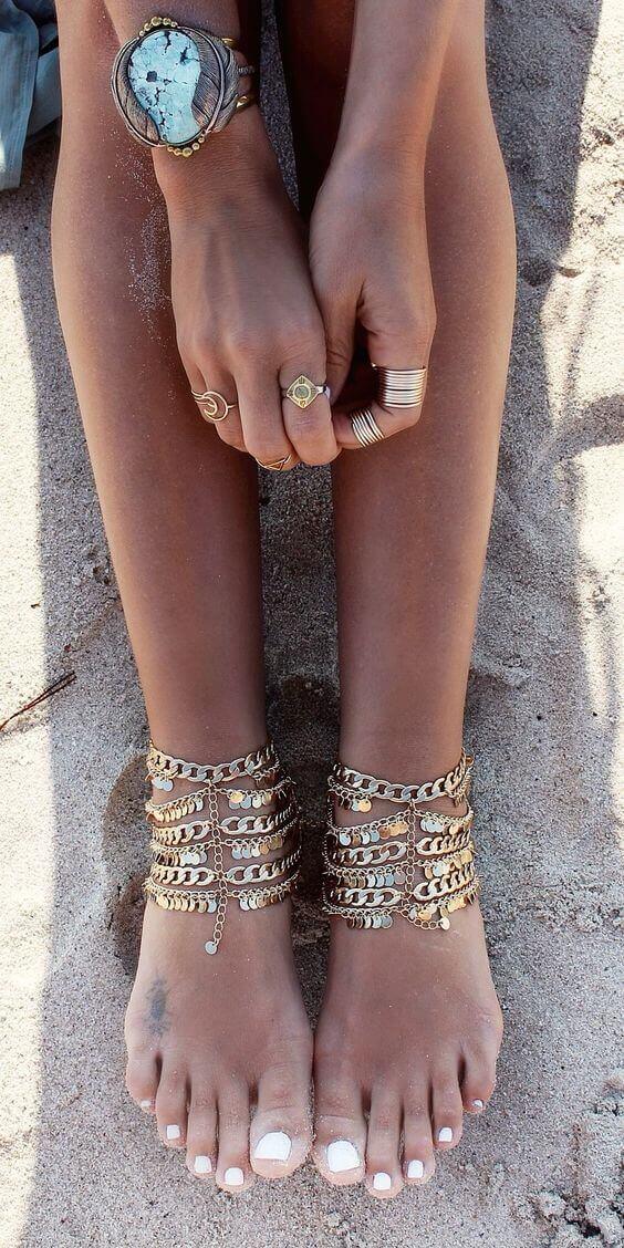 Anklets adorned with multiple chains and coins, complementing polished white nails, set against a sandy beach, embodying a bohemian vibe.